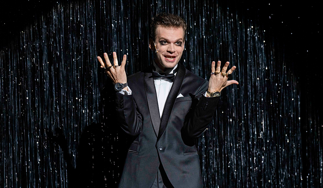 A man wearing a black suit and bow tie holds his hands up. He is wearing lots of large rings. The background is silver-spangled.